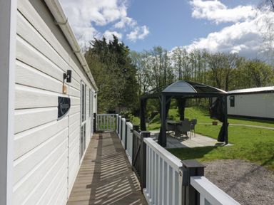 A white lodge with a wooden deck and railing next to a metal gazebo with chairs and table on a lawn at Ladybird Lodge South Lakes Leisure Park near Carnforth