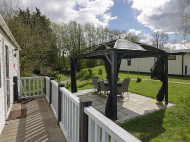A wooden deck with railing next to a gazebo with table and chairs on a stone patio at Ladybird Lodge South Lakes Leisure Park near Carnforth