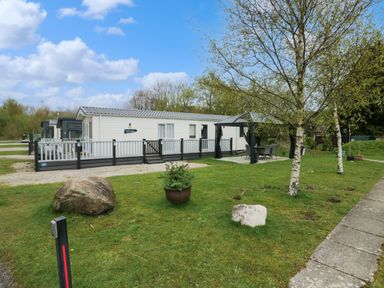 A white mobile home with a fenced porch and outdoor seating under a gazebo on a grassy lot with trees at Ladybird Lodge South Lakes Leisure Park near Carnforth