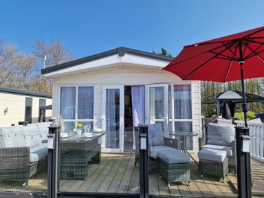 An outdoor patio with wicker furniture and a red umbrella in front of a white cabin at Ladybird Lodge South Lakes Leisure Park near Carnforth