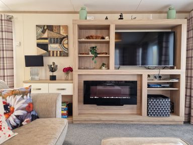 A living room with a TV and fireplace in a wooden unit next to a sofa with floral cushions at Ladybird Lodge South Lakes Leisure Park near Carnforth