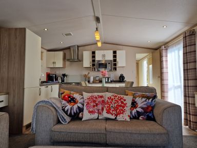 A living room with a sofa and decorative pillows facing a kitchen with cabinets and appliances at Ladybird Lodge in South Lakes Leisure Park near Carnforth