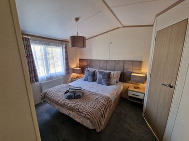 A bedroom with a bed with pillows and folded towels two bedside tables with lamps a window with curtains and a door at Ladybird Lodge South Lakes Leisure Park near Carnforth