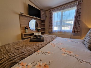 A bedroom with a bed covered in floral sheets and folded towels facing a wall-mounted tv and round mirror at Ladybird Lodge South Lakes Leisure Park near Carnforth