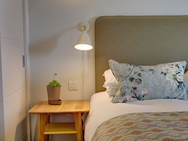 A bedside table with a potted plant next to a bed with a patterned pillow and a wall-mounted lamp at Villa 1 in Mullion