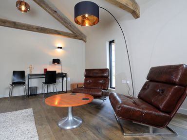 A living room with two leather chairs a round wooden table a desk with two black chairs and exposed ceiling beams at The Barns @ Downton in Dittisham