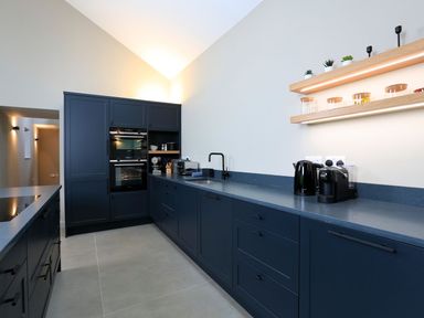 A kitchen with dark blue cabinets black appliances and wooden shelves with jars and plants at The Barns @ Downton in Dittisham