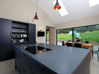 A kitchen with a large island and induction cooktop a dining table set near glass doors and cabinets with dishes at The Barns @ Downton in Dittisham