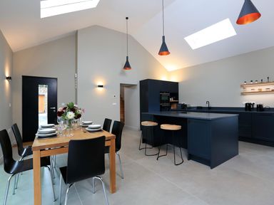 A kitchen with a dining table set with plates and glasses beside a black kitchen island with stools at The Barns @ Downton in Dittisham