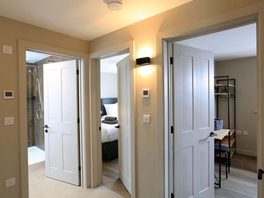 A hallway showing three open doors leading to a bathroom with a shower bedroom with a bed and towels and a room with a desk and chair at The Barns @ Downton in Dittisham
