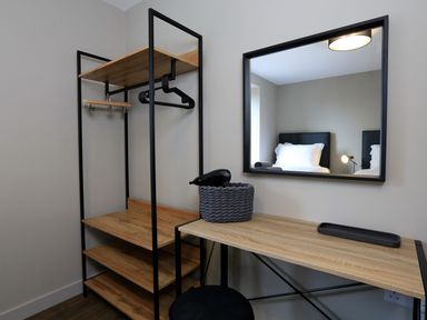 A wooden desk with a basket and hairdryer next to a shelving unit with hangers and a mirror reflecting a bed at The Barns @ Downton in Dittisham
