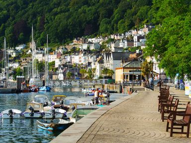 Harbor with boats docked along the quay and houses on a hillside near a walkway with benches at The Barns @ Downton in Dittisham