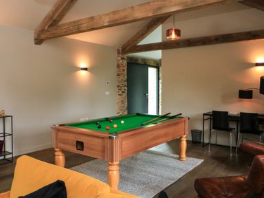 A room with a pool table on a rug next to a yellow sofa and desk with chairs at The Barns @ Downton in Dittisham