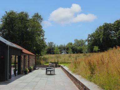 An outdoor patio area with chairs and a table next to a stone building and grassy fields at The Barns @ Downton in Dittisham