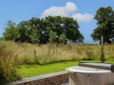 An outdoor hot tub next to grass and wild plants with trees in the background at The Barns @ Downton in Dittisham