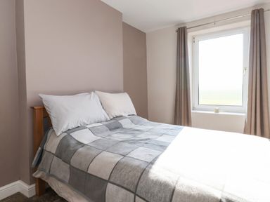 A bedroom with a double bed covered with a checkered blanket next to a window with curtains at 2 South Street in Fletchertown near Aspatria