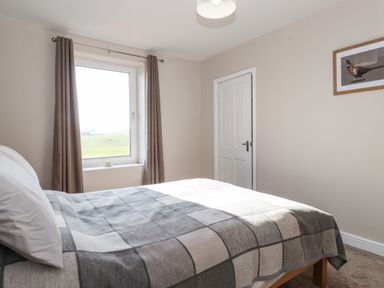 A bedroom with a bed covered in a checkered blanket near a window with curtains and a door on the right wall with a framed picture at 2 South Street in Fletchertown near Aspatria