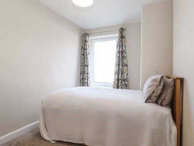 A bedroom with a bed covered in a white blanket and two pillows next to a window with floral curtains at 2 South Street in Fletchertown near Aspatria