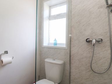 A bathroom with a toilet, toilet paper holder, shower with a handheld showerhead, and a window with a bottle on the sill at 2 South Street in Fletchertown near Aspatria