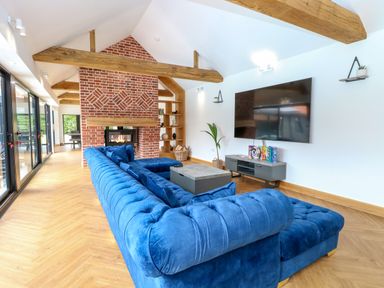 A living room with a blue sectional sofa a brick fireplace a large television and wooden beams at County Farm Barn in South Walsham