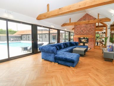 A living room with a blue sectional sofa a coffee table a brick fireplace wooden beams and large glass doors overlooking a pool at County Farm Barn in South Walsham
