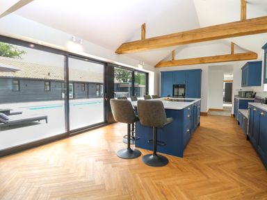 A kitchen with blue cabinets a large island with stools wooden beams on the ceiling and sliding glass doors at County Farm Barn in South Walsham