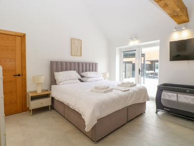 A bedroom with a large bed a nightstand with a lamp a wooden door glass doors leading outside a television and a baby cot at County Farm Barn in South Walsham
