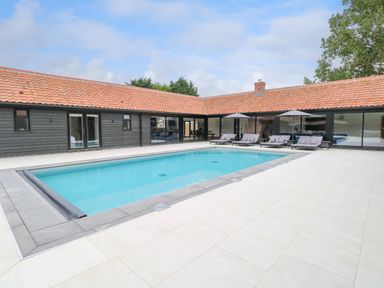 A rectangular swimming pool with lounge chairs and umbrellas outside a building with red tiled roof at County Farm Barn in South Walsham