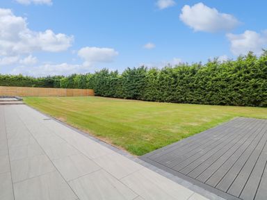 A backyard with a tiled patio a wooden deck a grass lawn and a fence with tall trees at County Farm Barn in South Walsham