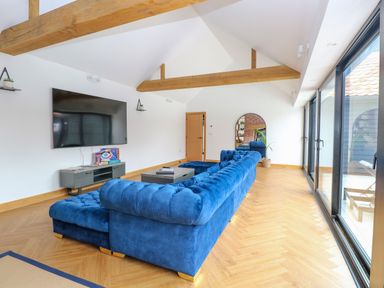 A living room with a blue sectional sofa a wall-mounted television wooden beams and large sliding glass doors at County Farm Barn in South Walsham