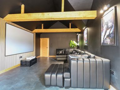 A media room with a large black sectional sofa a projector screen wooden beams on the ceiling and framed movie posters at County Farm Barn South Walsham
