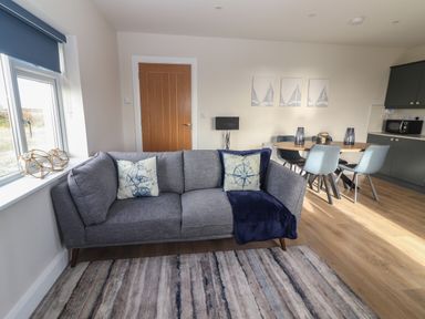 A living area with a gray sofa with pillows and blanket near a window and a dining table with chairs at The Boathouse in Carrigaholt County Clare