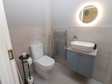 A bathroom with a toilet a wall-mounted sink with cabinet a circular illuminated mirror and a towel rack at The Boathouse in Carrigaholt County Clare