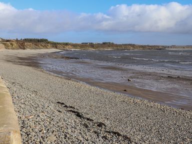 A pebbled beach with a low tide and distant cliffs under a partly cloudy blue sky at The Boathouse in Carrigaholt County Clare