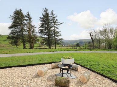 An outdoor fire pit with logs placed around it on gravel with trees and hills in the background at Willow in Llangurig near Llanidloes