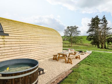 An outdoor picnic table and benches on gravel next to a curved wooden building with a hot tub and grassy field in the background at Willow in Llangurig near Llanidloes