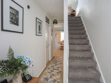 A hallway with a staircase carpeted in grey a wooden telephone table with flowers and framed pictures on the wall at Waters Edge Ymyl Y Dwr in Y Felinheli