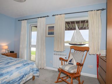 A bedroom with a bed bedside table with lamp rocking chair and window with curtains at Waters Edge Ymyl Y Dwr in Y Felinheli