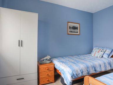 A bedroom with two single beds a wooden nightstand with a model boat a white wardrobe and a framed picture on a blue wall at Waters Edge Ymyl Y Dwr in Y Felinheli