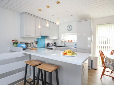 A kitchen with white cabinets a kitchen island with two stools and a fruit bowl at Waters Edge Ymyl Y Dwr in Y Felinheli