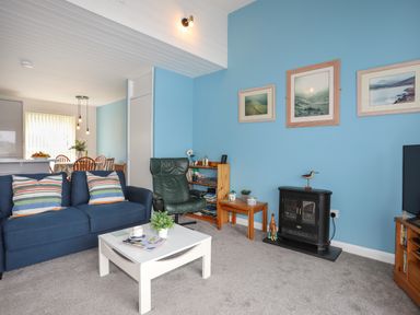 A living room with a blue sofa, green armchair, white coffee table, small fireplace, and framed pictures on a blue wall at Waters Edge Ymyl Y Dwr in Y Felinheli