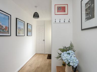 A hallway with framed pictures on the wall a white door light wood flooring and a vase with blue flowers at Waters Edge Ymyl Y Dwr in Y Felinheli