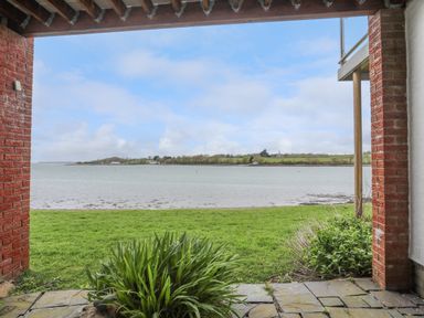 A covered patio opening to a grassy area by a body of water with land and houses in the distance at Waters Edge Ymyl Y Dwr in Y Felinheli