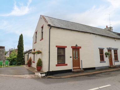 A street view of a connected two story and one story house with potted plants and hanging flower baskets at River Cottage in Eamont Bridge