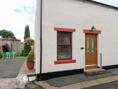 The exterior of a cottage with a wooden door and a window with red trim beside a small garden with green chairs at River Cottage in Eamont Bridge