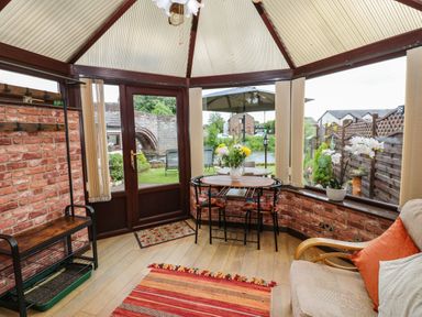 A conservatory with brick walls a small round table with four chairs a flowering plant and view of garden and stone bridge at River Cottage in Eamont Bridge