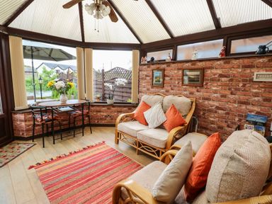A conservatory with wicker sofas with cushions a small dining table with chairs flowers in vases and a striped rug on the floor at River Cottage in Eamont Bridge