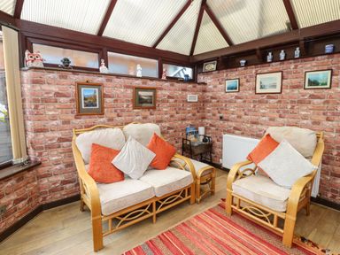A conservatory with rattan furniture cushions framed pictures on brick walls and a striped rug at River Cottage in Eamont Bridge