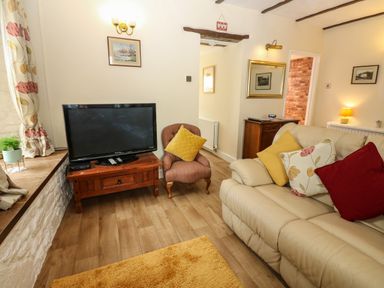 A living room with a beige sofa with cushions a wooden TV stand with a TV and a small armchair at River Cottage in Eamont Bridge
