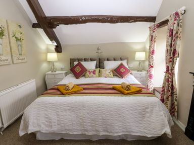 A bedroom with a large bed with striped runner and multiple pillows bedside tables with lamps floral curtains and wooden ceiling beams at River Cottage in Eamont Bridge